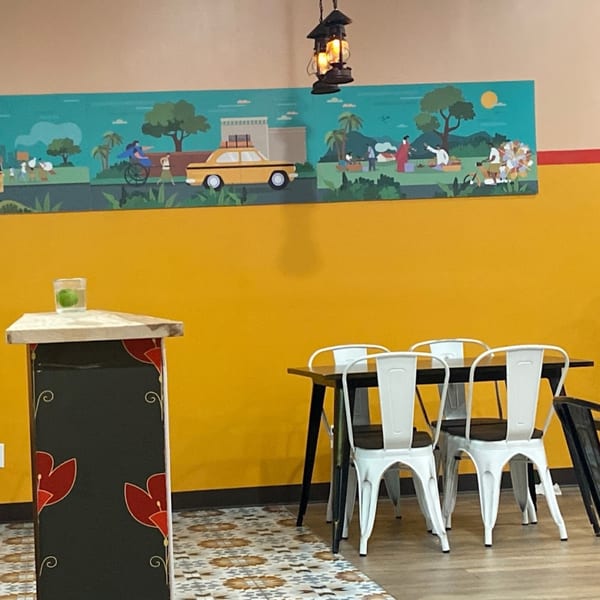 A vibrant Indian-themed mural above a table with white chairs and a decorative stand at Bittoo's Tikka.