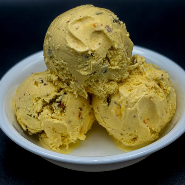 Three scoops of golden Kulfi ice cream with nuts, served in a white bowl.