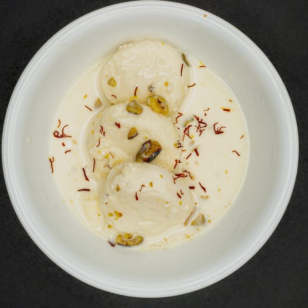 Three pieces of Ras Malai, an Indian dessert, in a white bowl, garnished with saffron and pistachios.