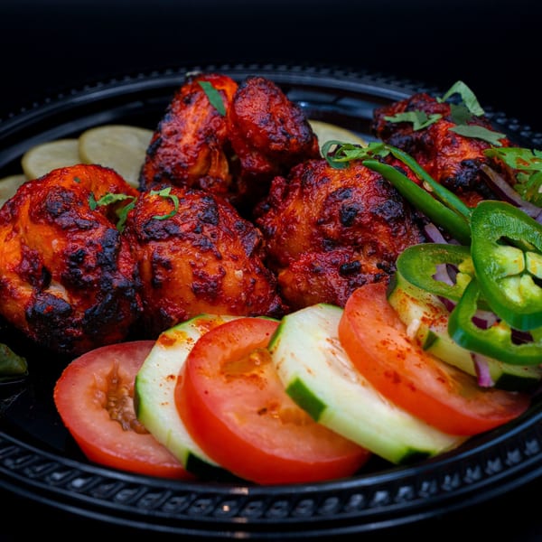 Bittoo's Tikka chicken tikka with sliced tomatoes, cucumbers, and green chilies on a black plate.